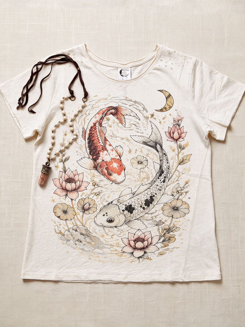 An off-white short-sleeve t-shirt with a detailed graphic print of two koi fish in a circular floral arrangement, styled flat on a beige linen background with a beaded necklace draped over the shoulder.