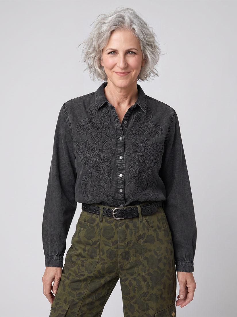 A woman with short gray hair wearing a dark charcoal gray long-sleeve button-up shirt by maison hotel with a subtle paisley pattern and olive green camouflage pants by maison hotel, standing against a light gray studio background.