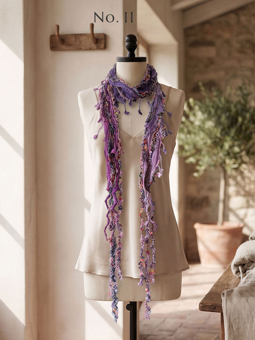 A long, fringed scarf by monkey mango in shades of purple and lavender displayed on a cream-colored dress form mannequin, set in a warm, sunlit room with a rustic stone wall and potted plant in the background.