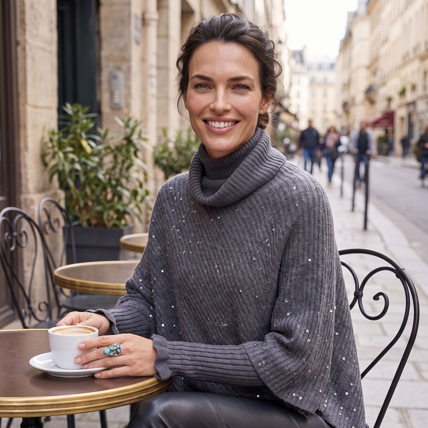 A smiling woman sits at an outdoor cafe table wearing a dark gray ribbed turtleneck sweater with subtle silver speckles. She holds a cup of coffee in her hand, with a cobblestone street and buildings visible in the background.