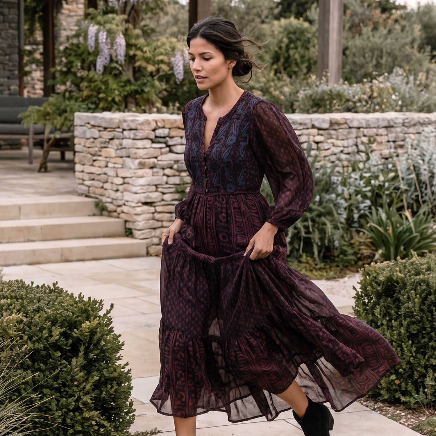 A woman wearing a dark burgundy midi dress with a tonal geometric pattern walks along a stone patio in a garden setting.