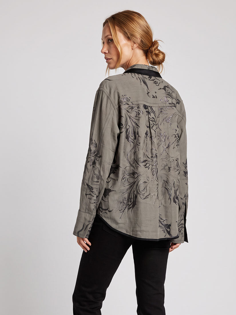 A woman shown from the back wearing a long-sleeve button-up shirt in gray with a black floral pattern, paired with black pants, against a light gray studio background.