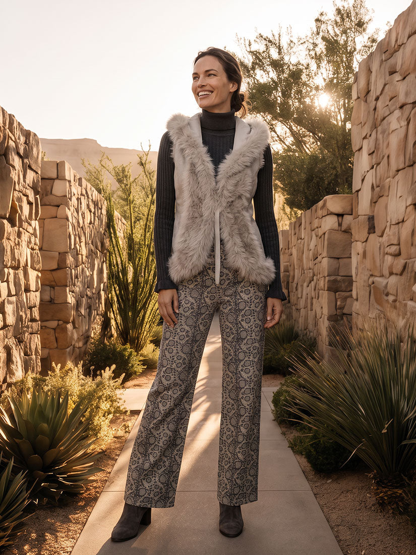 A woman stands on a stone walkway outdoors between stone walls and desert plants, wearing a light gray faux-fur vest over a dark ribbed turtleneck and patterned gray wide-leg pants.