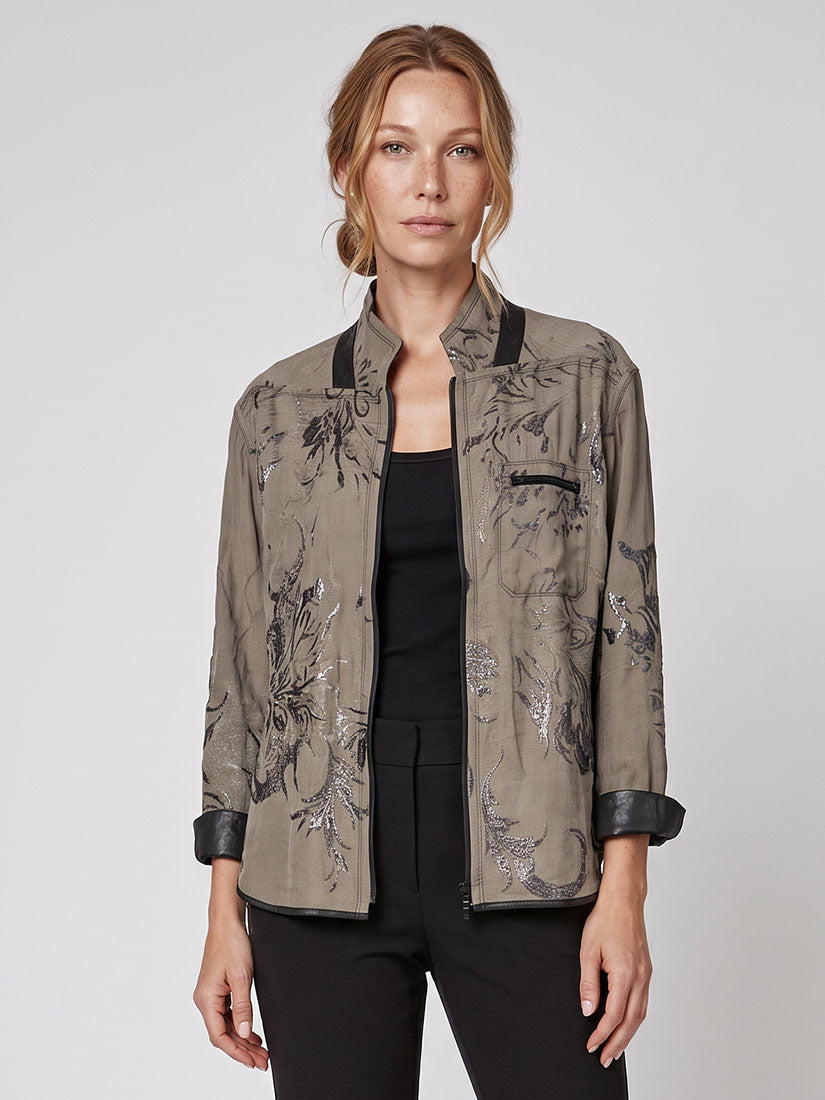 A woman modeling a taupe-gray patterned jacket with black trim and black sleeves, worn over a black top and black pants, against a light gray studio background.