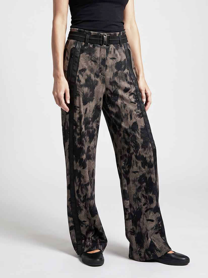 A model wearing high-waisted, wide-leg trousers with a black and brown abstract pattern, a black belt, and black flats, photographed against a light gray studio background.