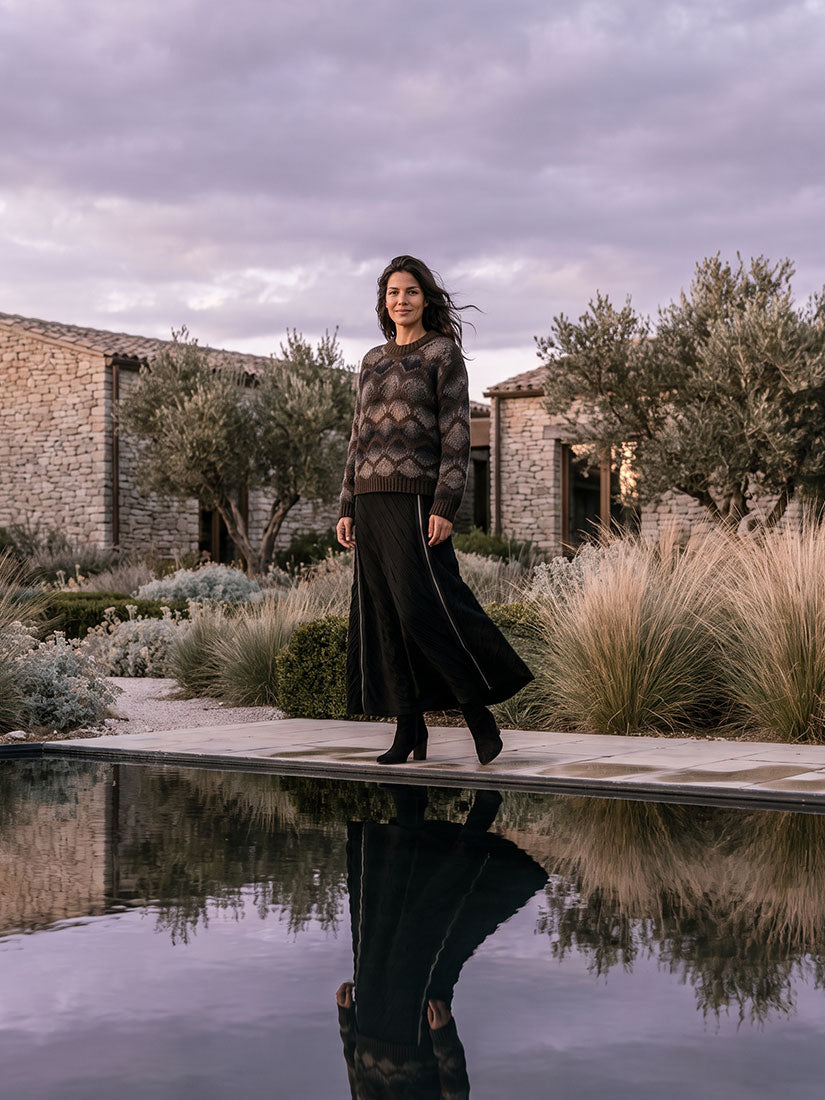 A woman stands outdoors near a reflective pool, wearing a dark patterned knit sweater and a long black skirt. Stone buildings, shrubs, and trees are visible in the background under a cloudy sky.