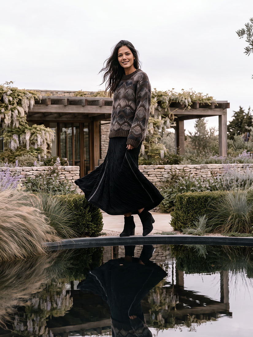 dark haired woman in black skirt and chunky knit sweater stand at edge of reflecting pool