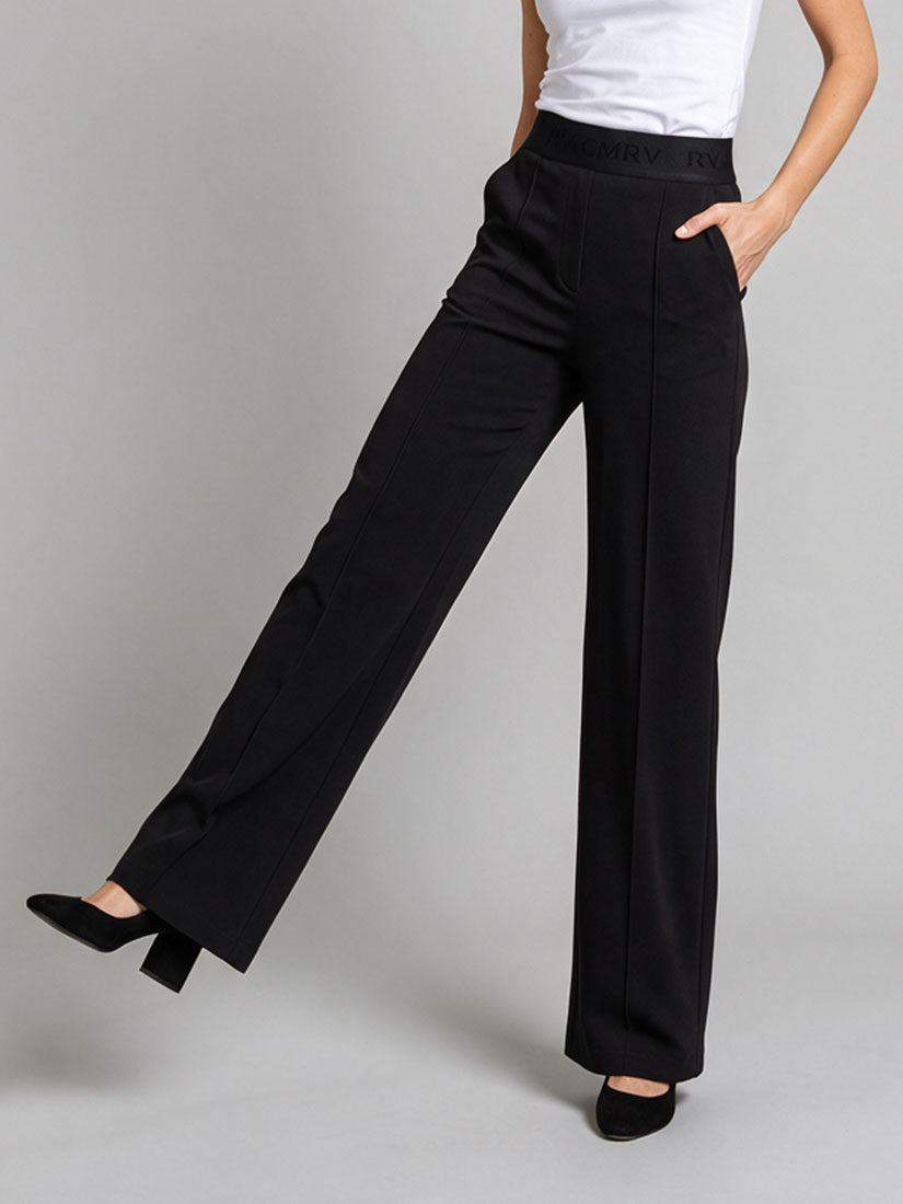 A model wearing black wide-leg trousers with a white top and black heels, shown from the waist down against a neutral gray studio background.