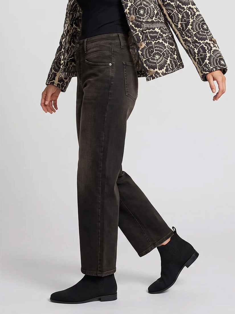 A model wearing a pair of dark brown straight-leg jeans, styled with a black top, a patterned jacket, and black suede ankle boots, shown from a side profile against a light gray studio background.