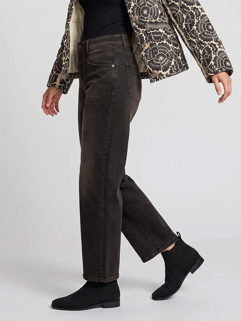A model wearing a pair of dark brown straight-leg jeans, styled with a black top, a patterned jacket, and black suede ankle boots, shown from a side profile against a light gray studio background.