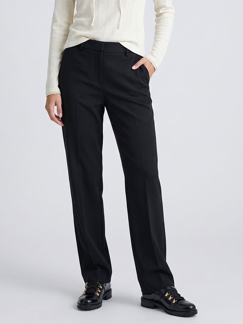 A model wearing black straight-leg trousers, a cream ribbed long-sleeve top, and black lace-up shoes with gold accents, standing against a light gray studio background.