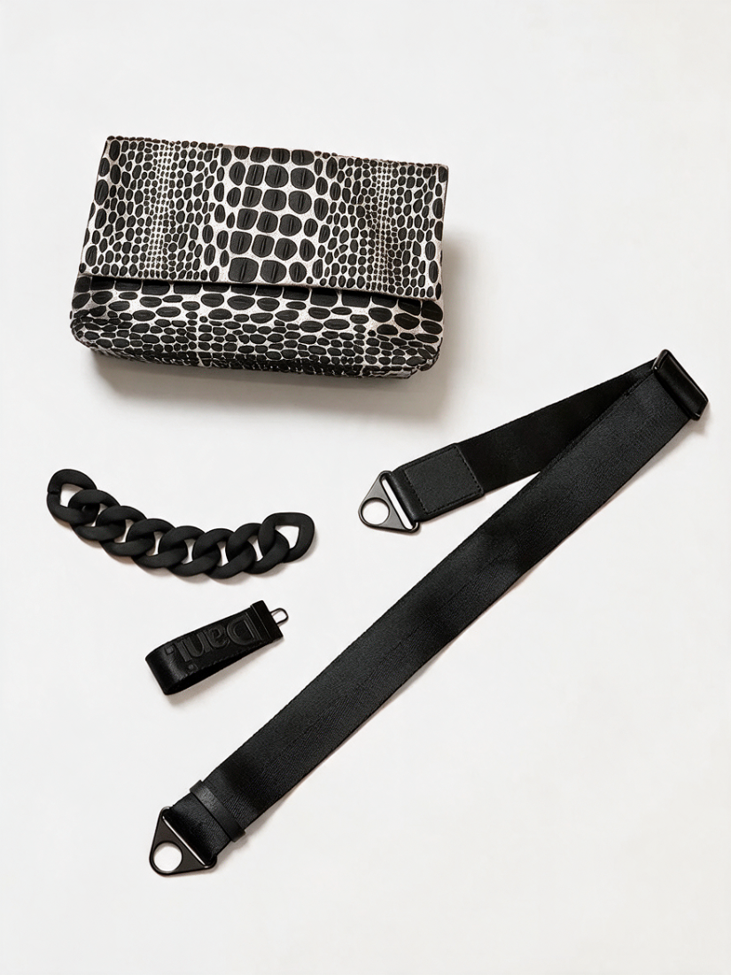 A black and metallic silver snakeskin-patterned clutch bag shown with a long black shoulder strap, a chunky black chain detail, and a small black strap section on a plain light background.