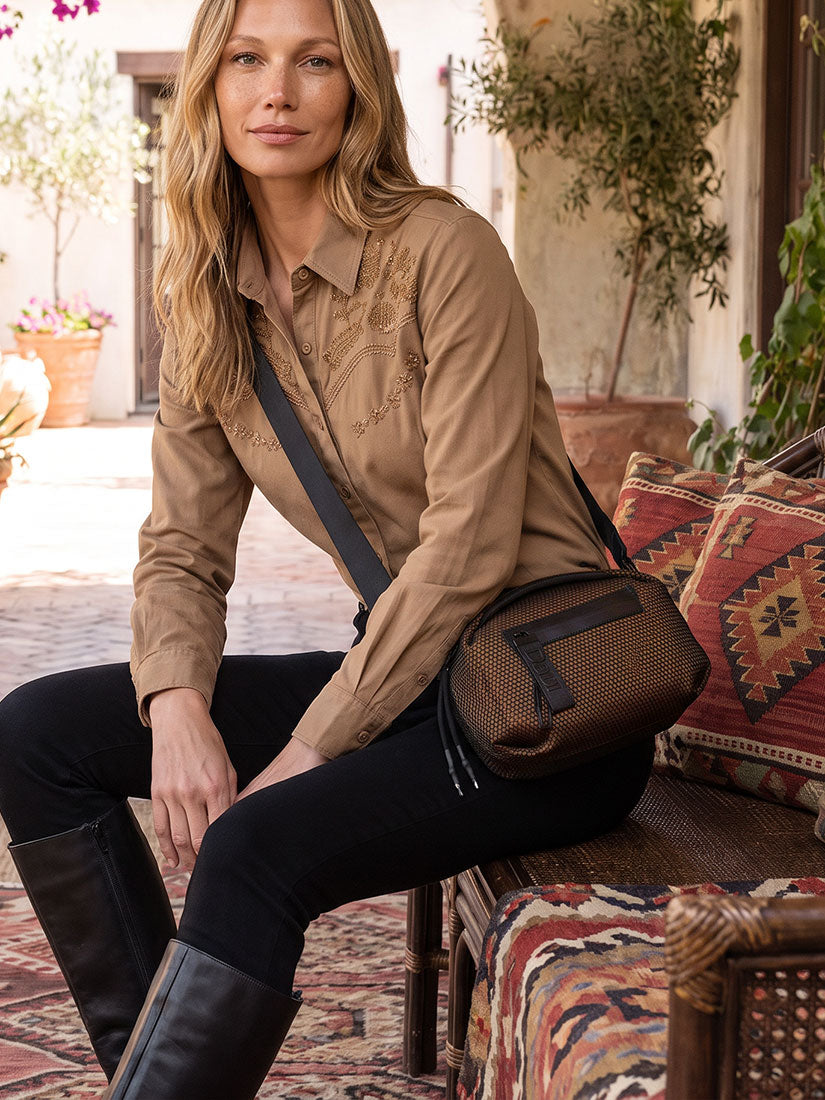 A woman sits on a patterned bench wearing a tan embroidered shirt, black pants, and black boots, with a brown crossbody bag slung across her body in a sunlit outdoor patio setting.