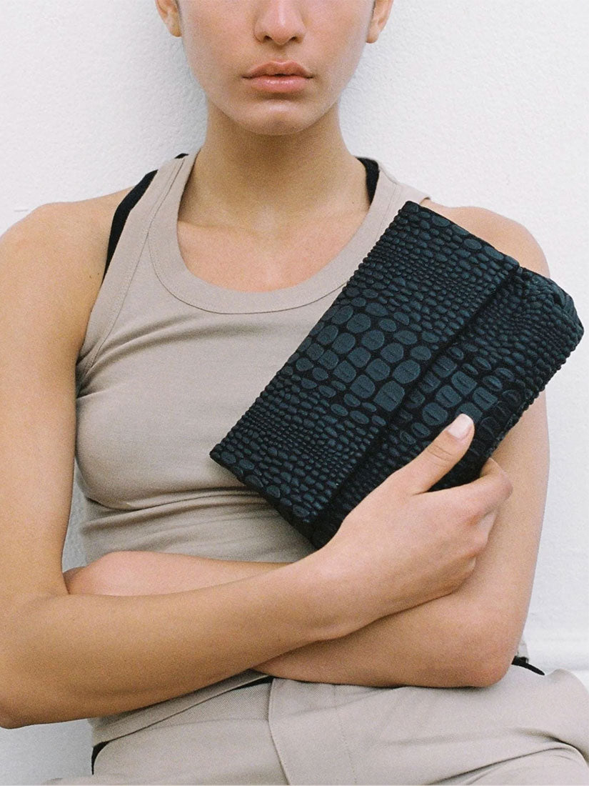 A model wearing a beige tank top holds a black tokyo crocodile-embossed clutch from daniella lehavi against her chest.