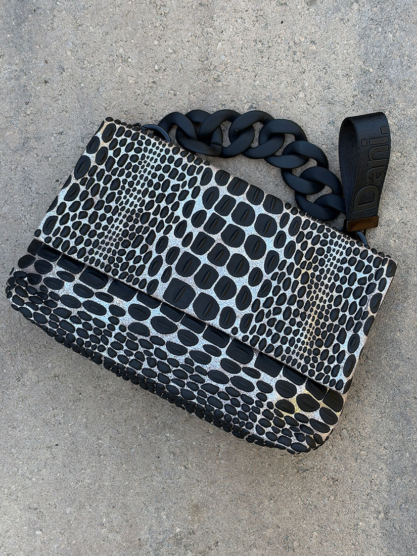 A black and metallic silver snakeskin-patterned clutch bag with a chunky black chain strap, laid flat on a gray concrete surface.