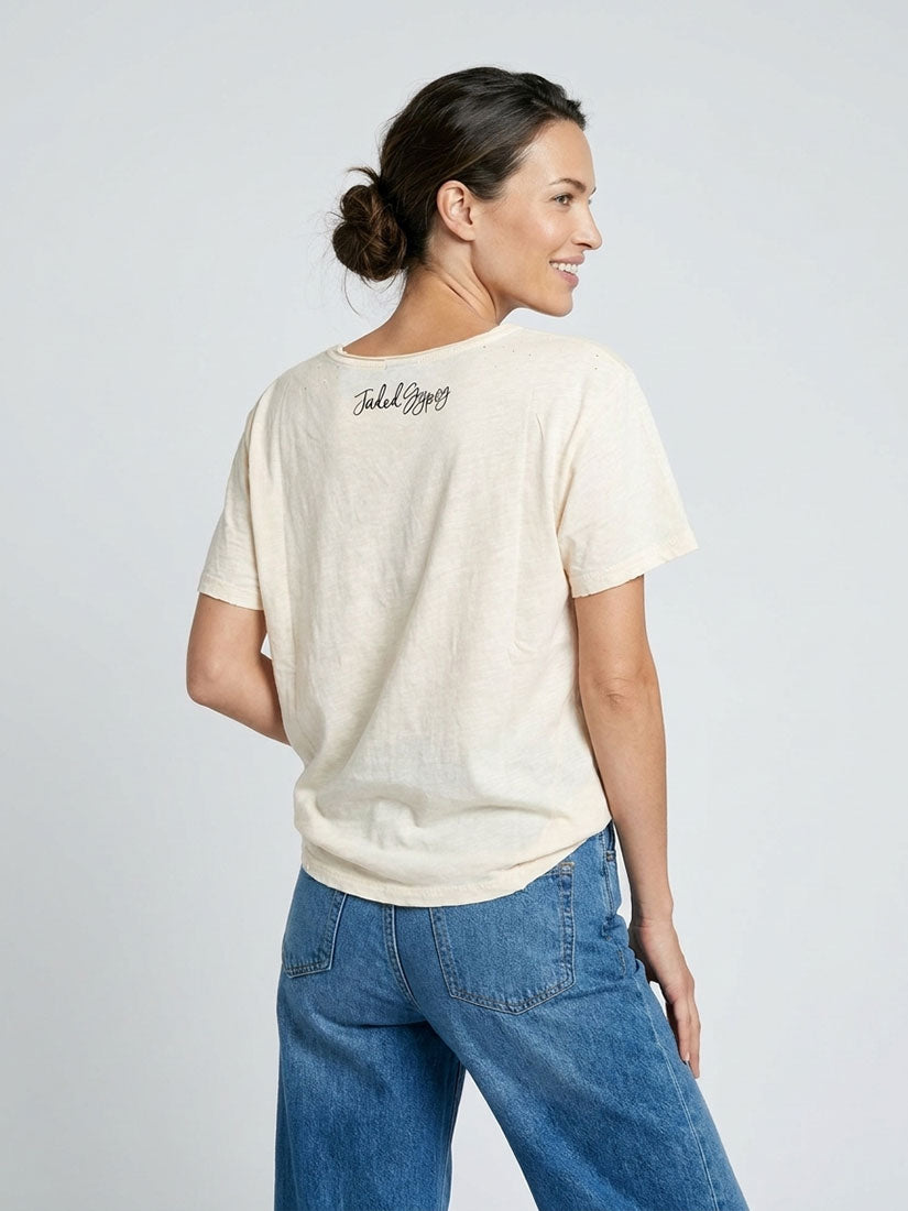 A woman wearing a cream-colored short-sleeve t-shirt with a small black script logo on the back, paired with blue jeans, photographed against a light gray studio background.