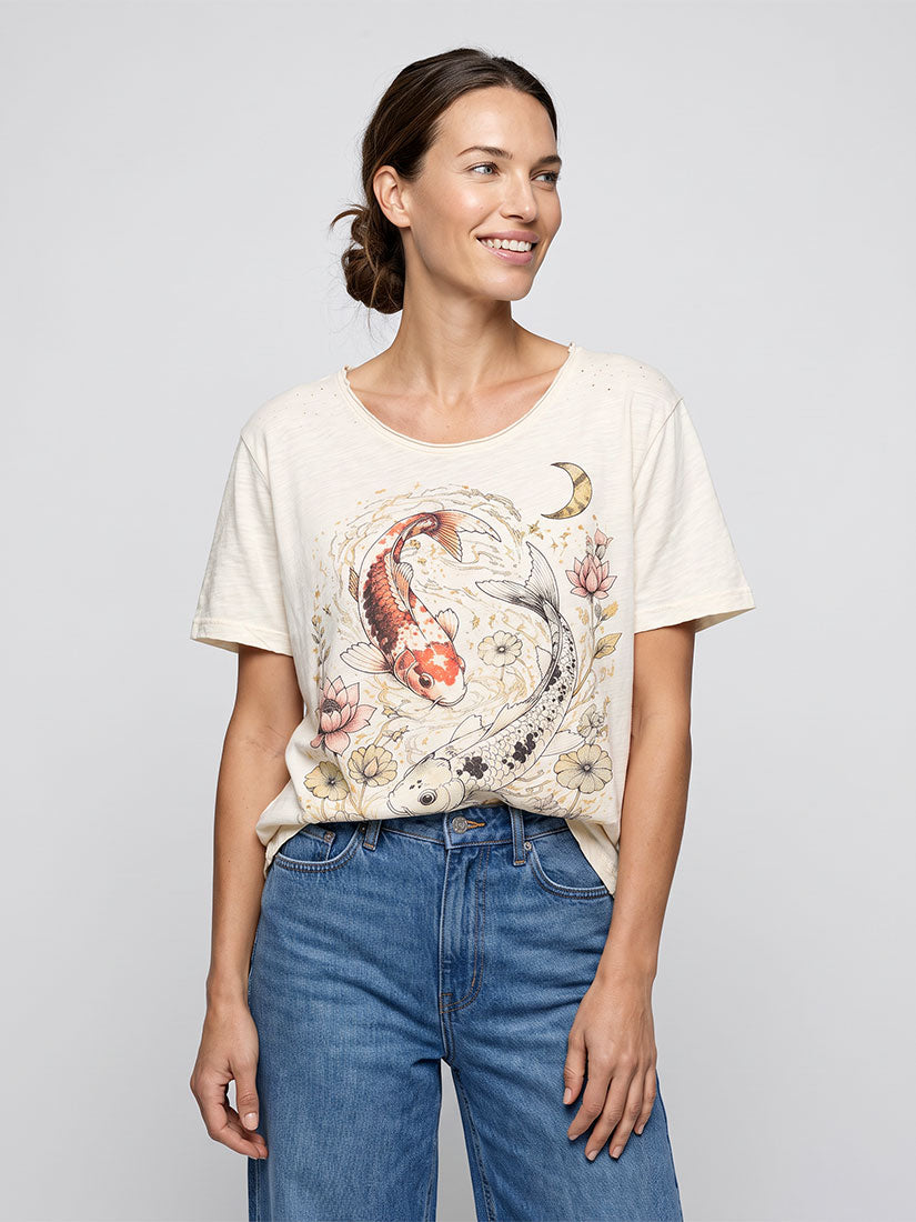 A woman modeling a cream-colored short-sleeve t-shirt with a detailed koi fish and floral graphic print, paired with blue jeans, against a solid light gray background.