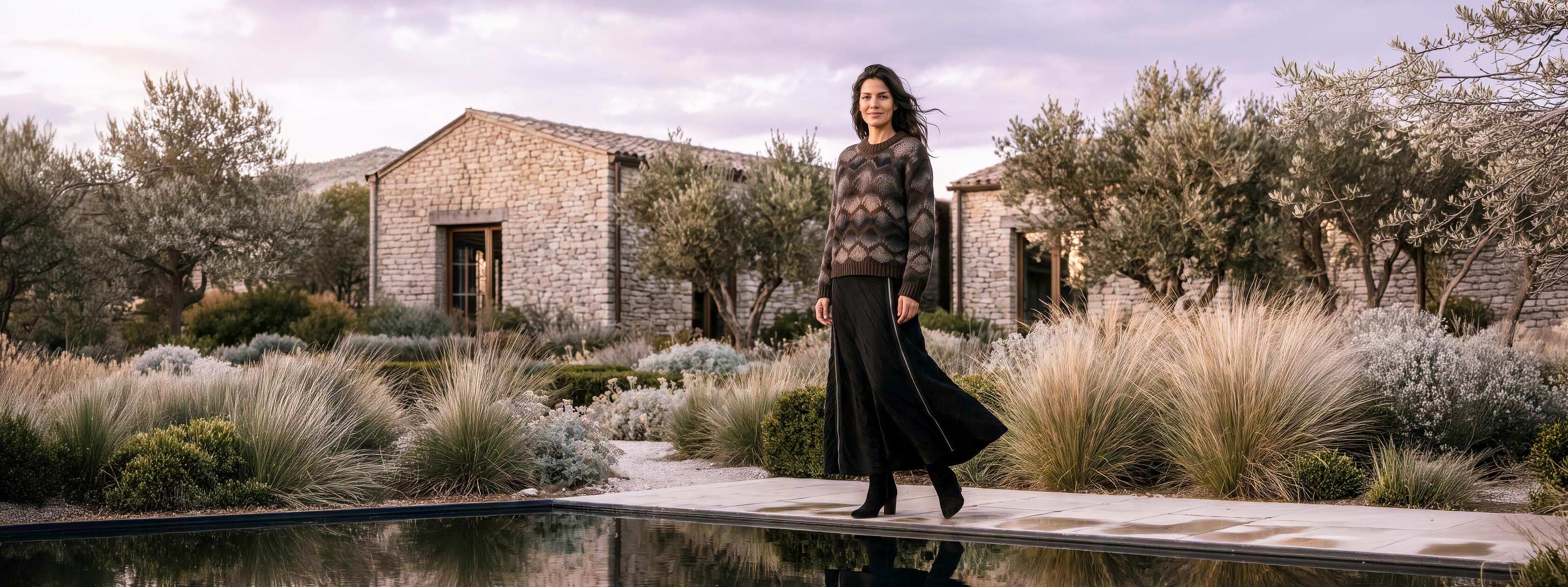 A woman stands outdoors in front of rustic stone buildings and olive trees, wearing a dark patterned knit sweater and a long black skirt. A reflective pool and ornamental grasses are visible in the foreground.