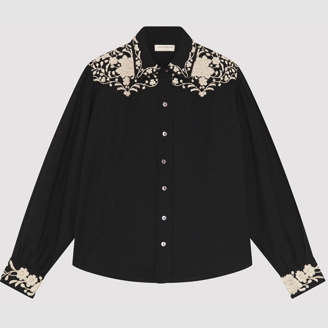 A black long-sleeve button-up shirt with intricate beige floral embroidery on the collar and cuffs, displayed flat against a light gray background.
