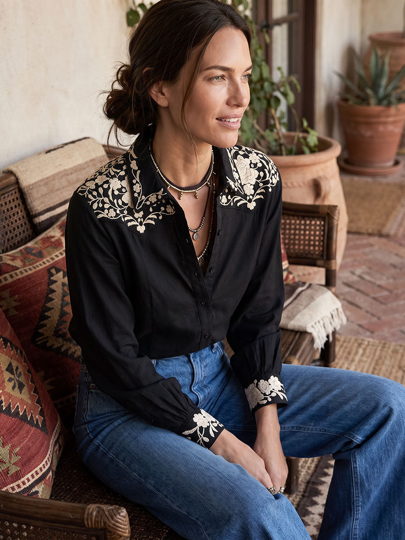 A woman sits on a woven chair wearing a maison hotel black long-sleeve antonio blouse with ornate white floral embroidery on the shoulders and cuffs, paired with blue jeans in a rustic indoor setting.