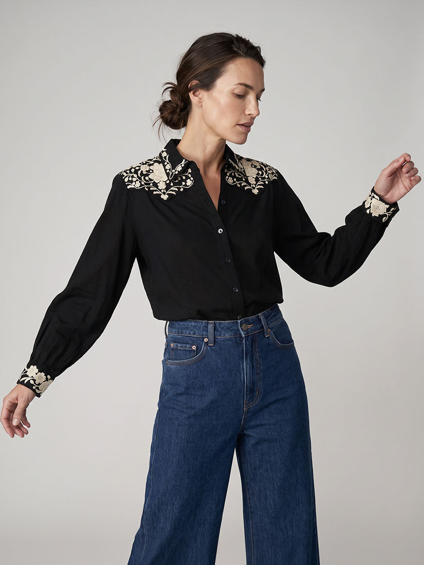 A woman modeling a maison hotel antonio black long-sleeve button-up blouse with ornate beige floral embroidery on the shoulders and cuffs, paired with high-waisted blue wide-leg jeans, against a neutral light gray studio background.