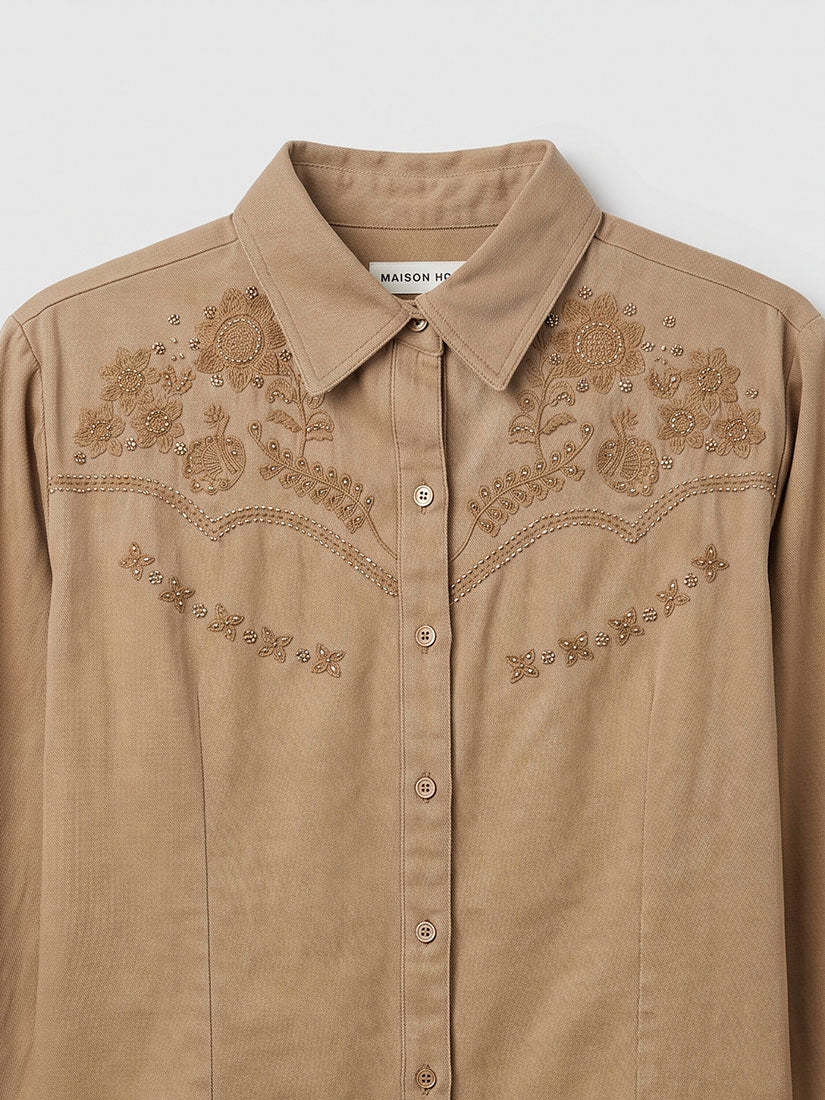 A tan western-style button-up shirt with intricate floral embroidery across the chest and shoulders, displayed on a neutral light gray background.