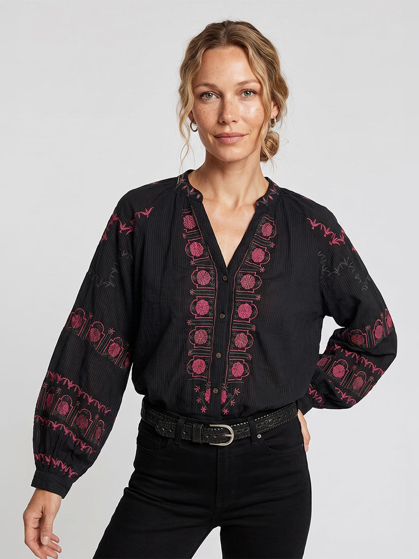 A woman modeling a black long-sleeve blouse with pink and red embroidered patterns on the front and sleeves, paired with black pants against a light gray studio background.