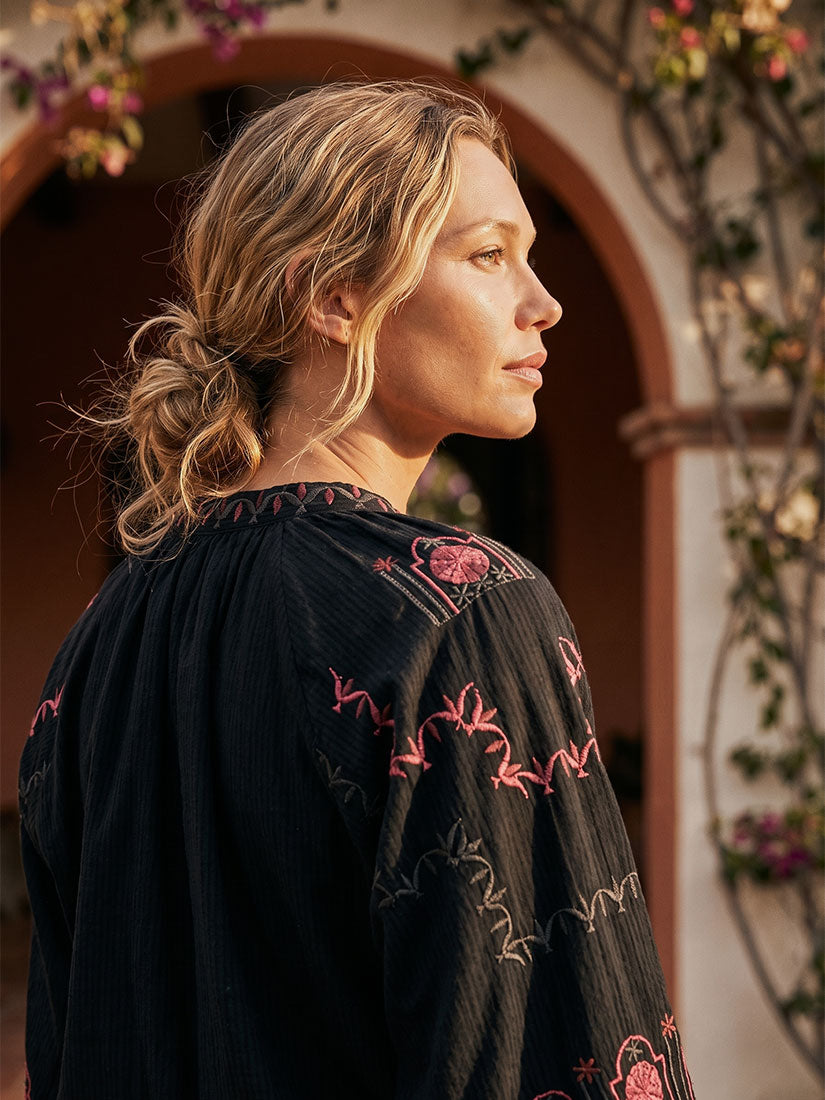 A woman in profile wearing a black textured blouse with pink floral embroidery, set against a warm, sunlit outdoor background with an archway and climbing vines.