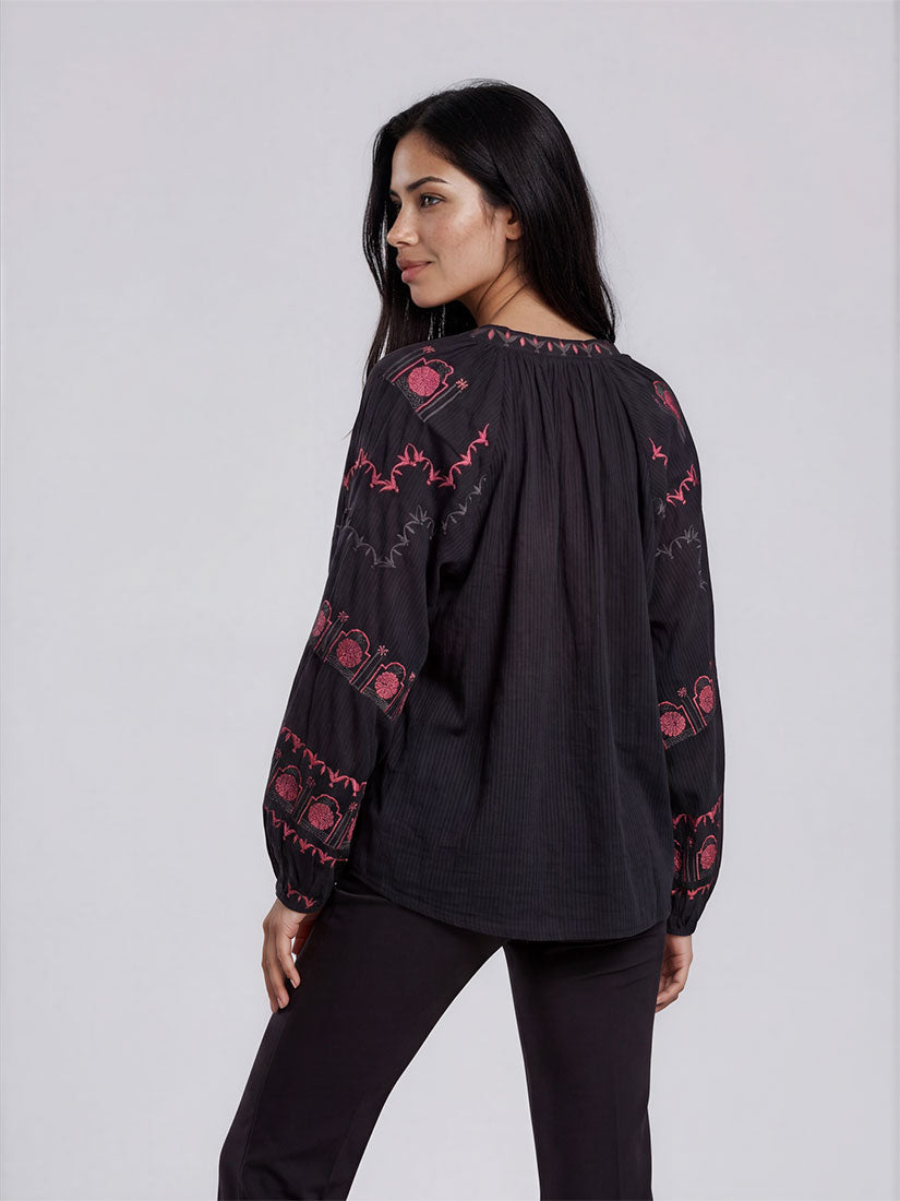 A woman wearing a black long-sleeve blouse with pink and red geometric embroidery, shown from a three-quarter back view against a light gray studio background.