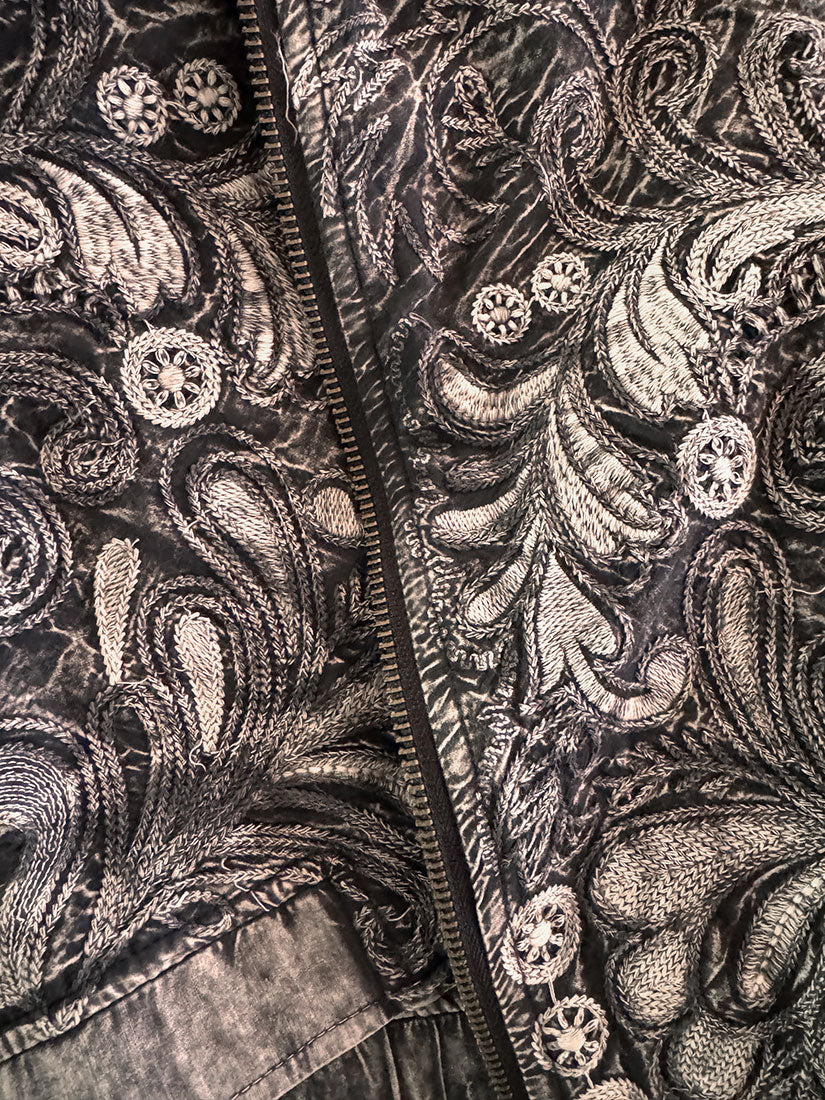 Close-up of a black and gray paisley embroidered jacket with a central zipper and decorative button details.