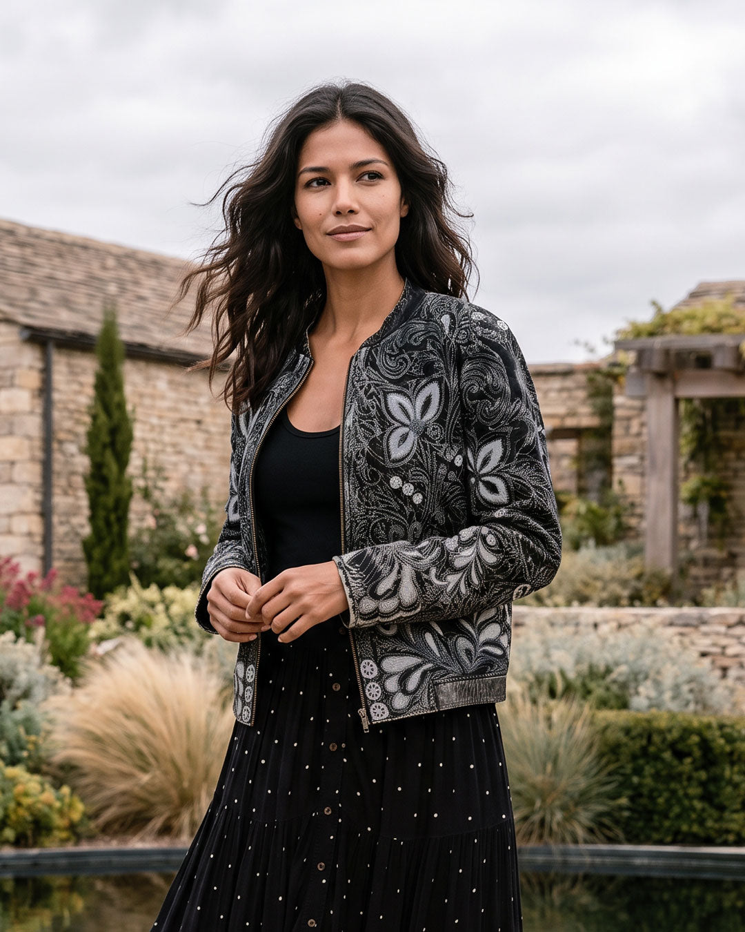 A woman stands outdoors in a garden courtyard wearing a black and gray embroidered bomber jacket over a black top and black polka-dot skirt.