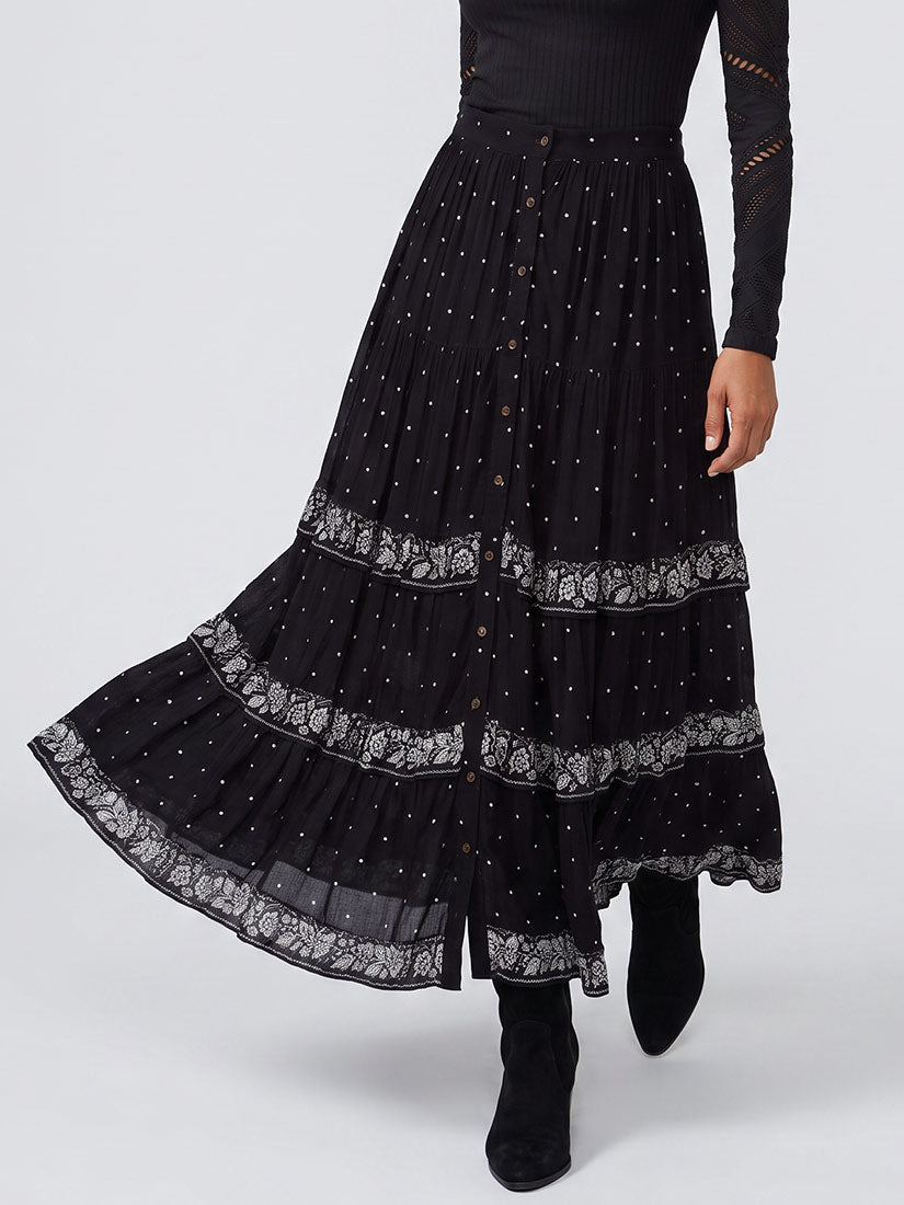 A black tiered maxi skirt with white lace trim and small white dot patterns, shown on a model wearing black ankle boots against a light gray studio background.