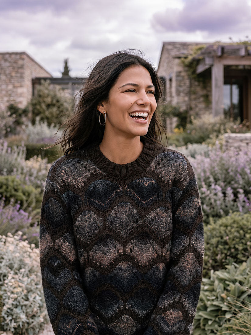 A woman smiling outdoors in front of stone buildings and garden plants, wearing a dark patterned knit sweater with a crew neck.