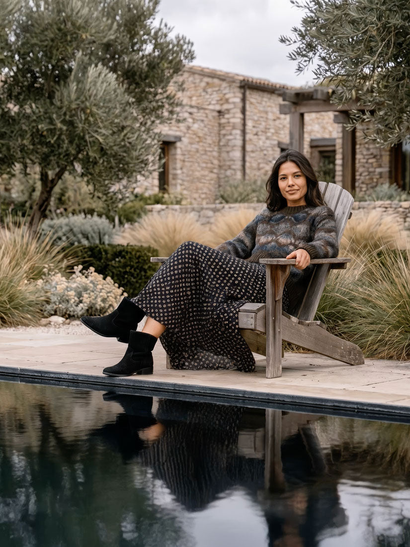 A woman sits in a wooden Adirondack chair beside a swimming pool in a stone courtyard garden. She is wearing a long-sleeve patterned knit top and a long black dress with small white dots, paired with black ankle boots.