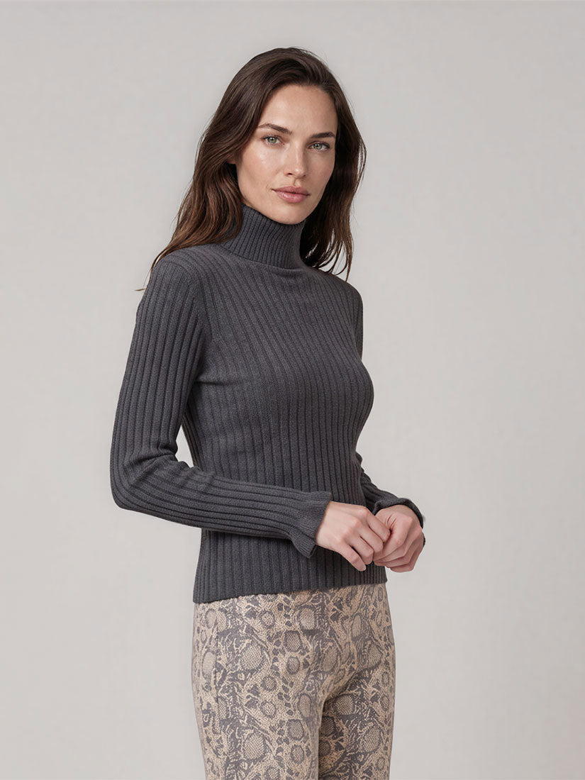 A woman modeling a dark gray ribbed turtleneck sweater paired with beige patterned pants, photographed against a neutral light gray studio background.
