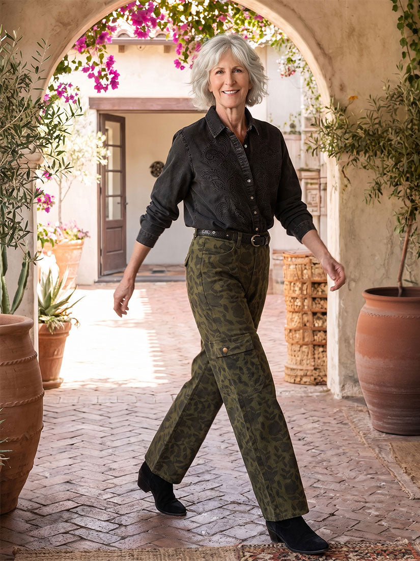 A smiling woman with gray hair walks through a sunlit stone courtyard framed by an archway. She is wearing a dark charcoal button-up shirt and olive green camouflage trousers by maison hotel, paired with black shoes.