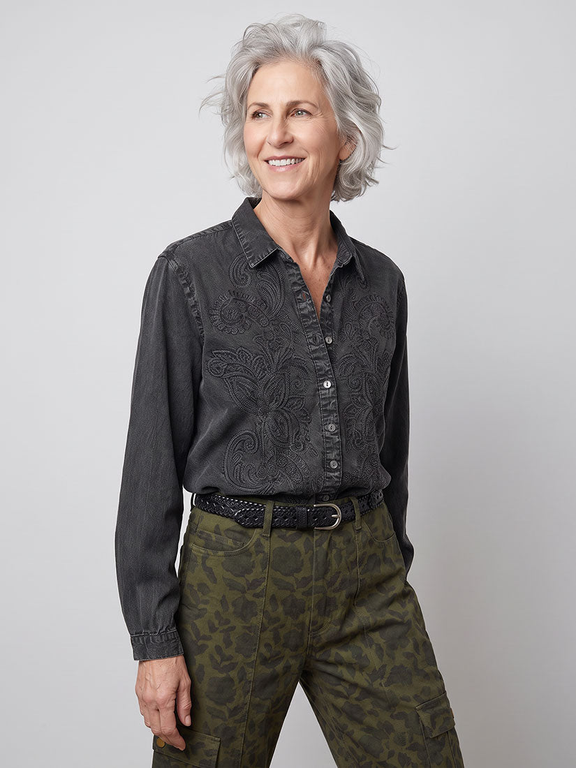 A smiling older woman with short gray hair is shown from the waist up against a light gray studio background. She is wearing a petra dark charcoal gray long-sleeve button-up shirt with a subtle tonal floral or paisley embossed pattern by maison hotel, paired with olive green patterned pants and a black belt.