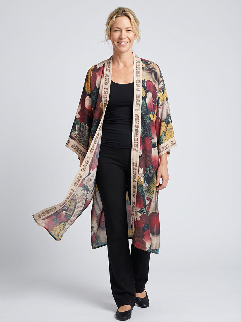 A woman modeling a lightweight open-front kimono with a busy multicolor floral and botanical print, worn over a black outfit against a light gray studio background.