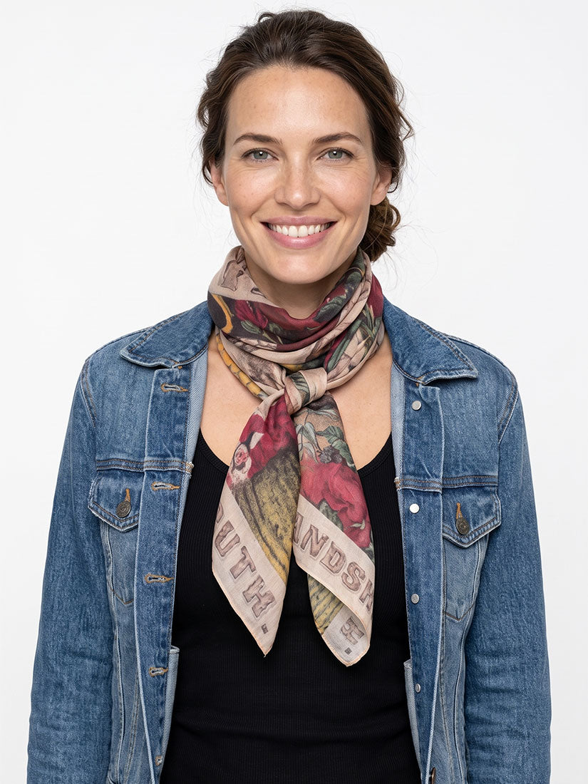 A woman wearing a patterned silk scarf with vintage-style illustrations and text, styled around her neck with a blue denim jacket over a black top, against a plain white background.