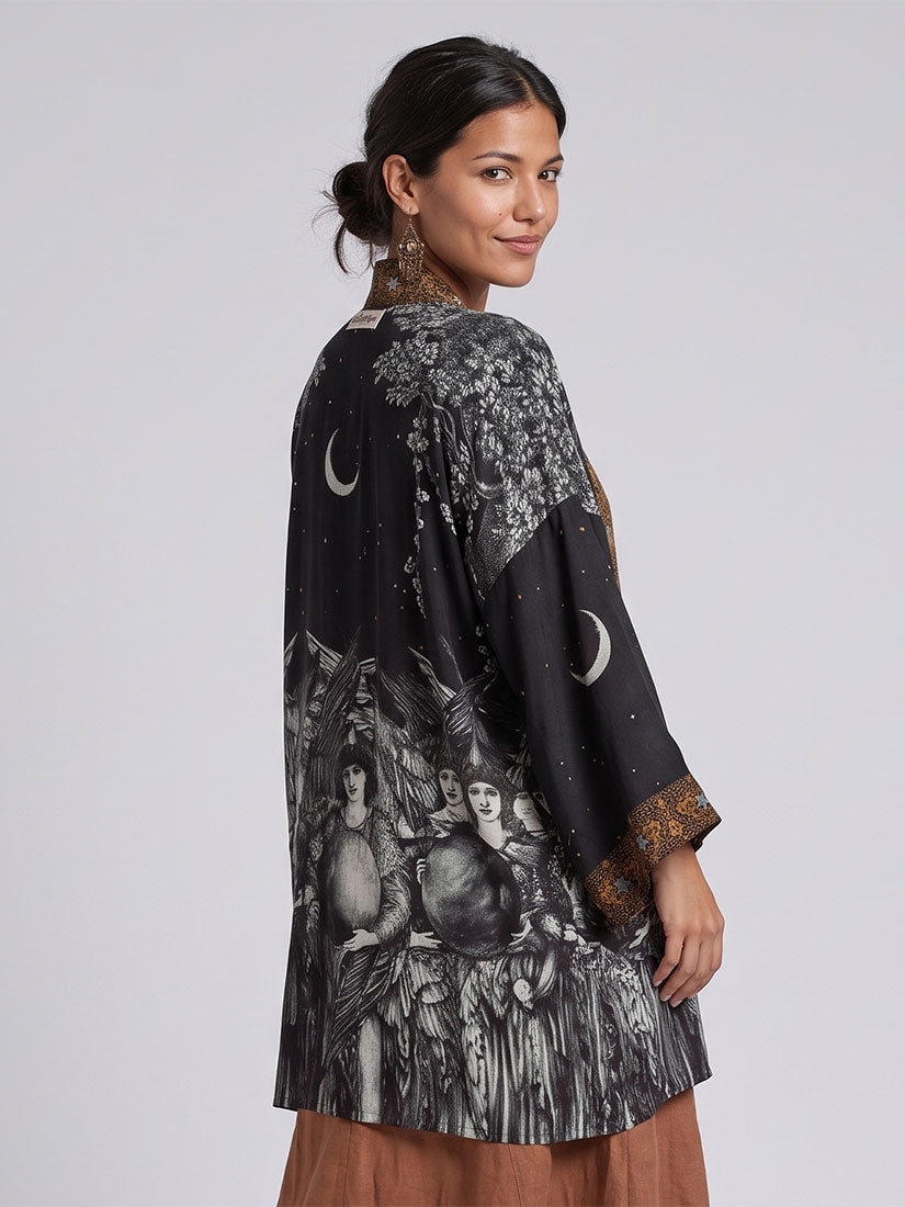 A woman modeling a black long-sleeve tunic with a celestial and gothic illustration print, featuring crescent moons, stars, and figures, paired with brown wide-leg pants against a light gray studio background.