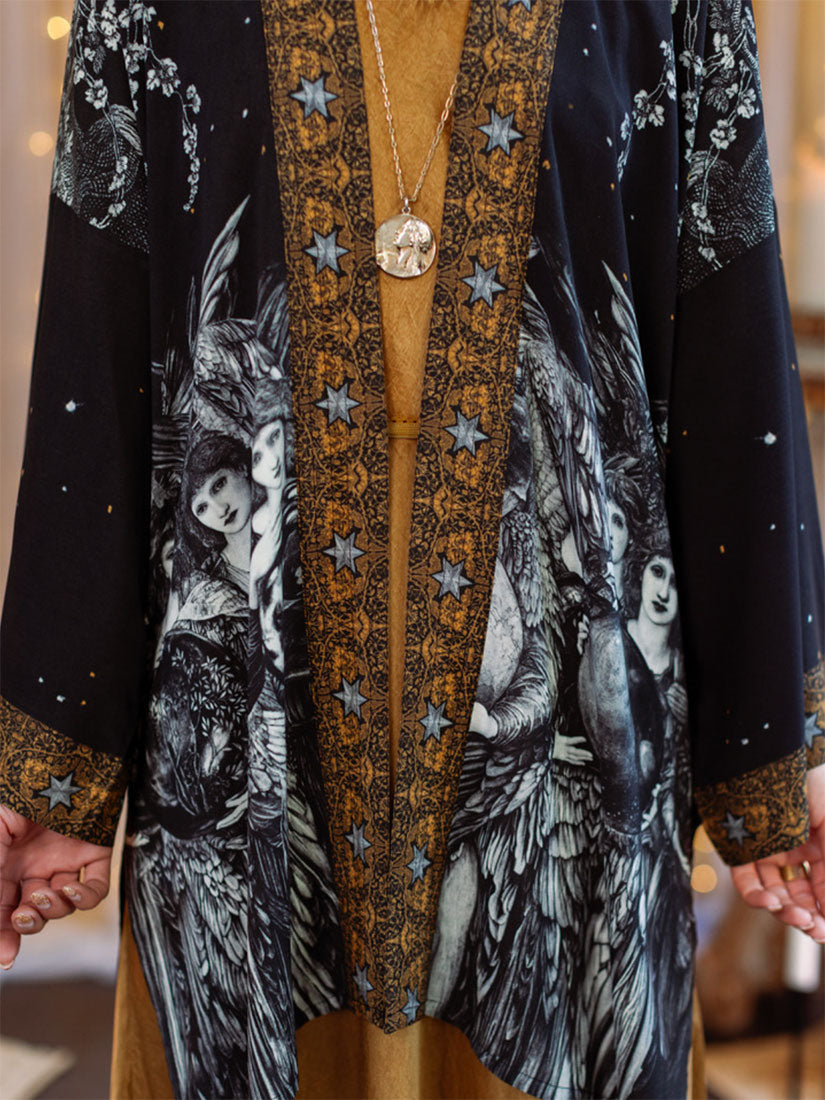 A close-up of a black open-front cardigan with a gold patterned center panel and star motifs, featuring black-and-white angel illustrations on the sleeves and front, worn over a tan dress.