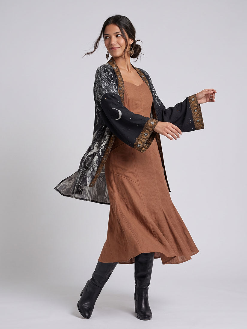 A woman models a bohemian-style outfit featuring a brown linen dress, a black embroidered kimono jacket with a moon motif, and black leather boots, set against a plain light gray studio background.