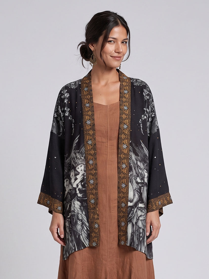 A woman modeling a black open-front kimono-style cardigan with a gray and white animal print and gold patterned trim, worn over a brown dress against a light gray studio background.