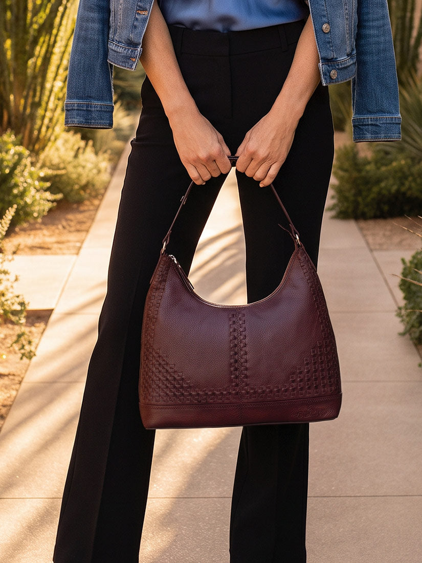 A person outdoors holding a burgundy leather hobo shoulder bag with woven texture details. The person is wearing black pants, a blue top, and a denim jacket, standing on a paved walkway with plants in the background.
