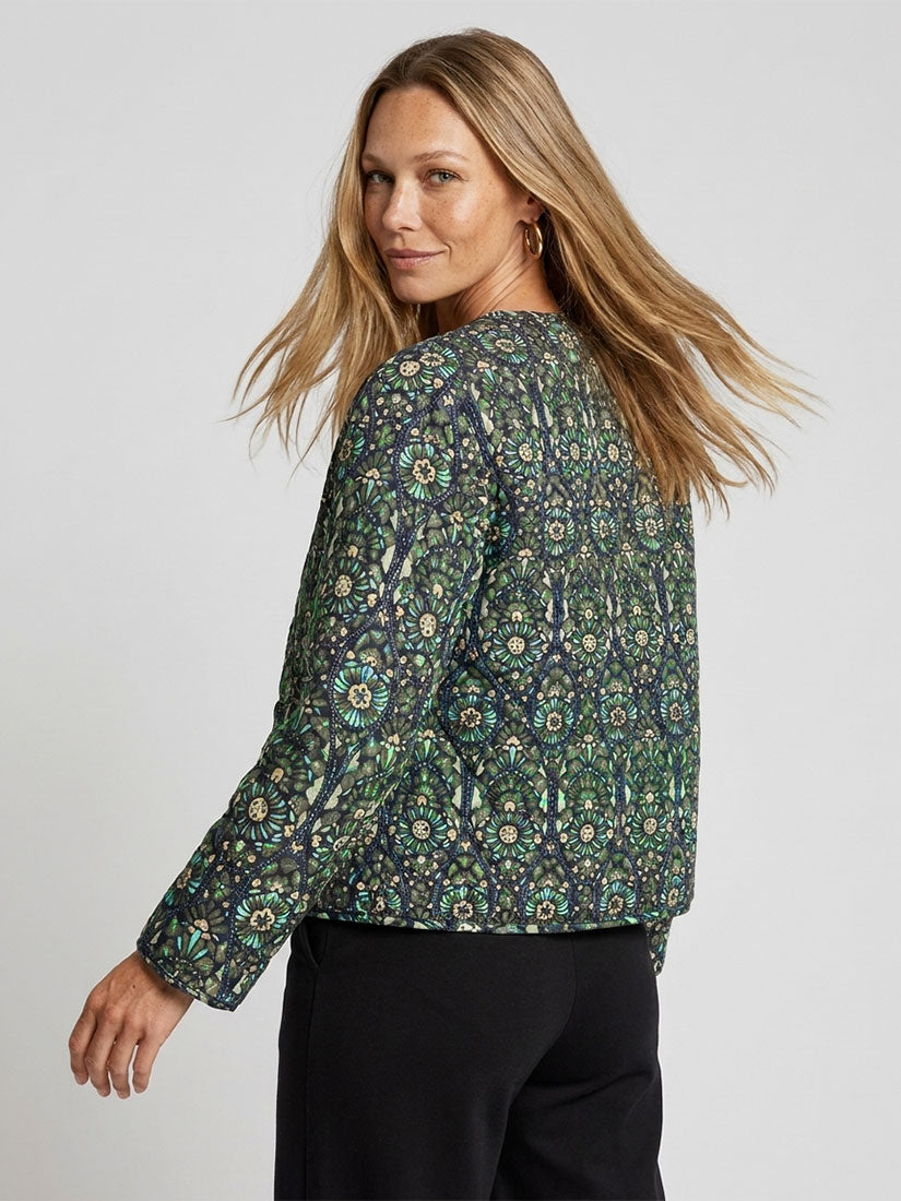A woman modeling a long-sleeve cropped jacket with an intricate green, blue, and beige paisley pattern, paired with black trousers, photographed against a solid light gray background.
