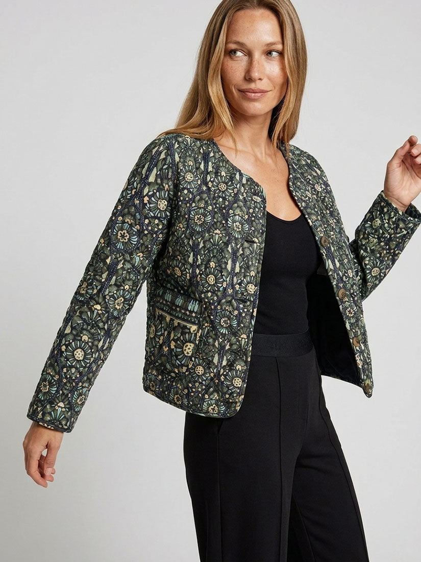 A woman modeling a cropped, long-sleeve quilted jacket with an intricate floral and botanical pattern in olive green, black, and beige, worn over a black top and black trousers against a light gray studio background.