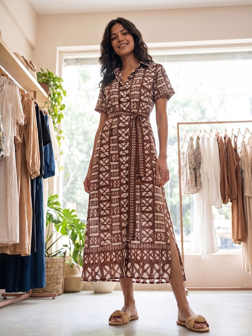 A woman modeling a brown and beige patterned midi dress in a boutique setting with clothing racks and plants in the background.