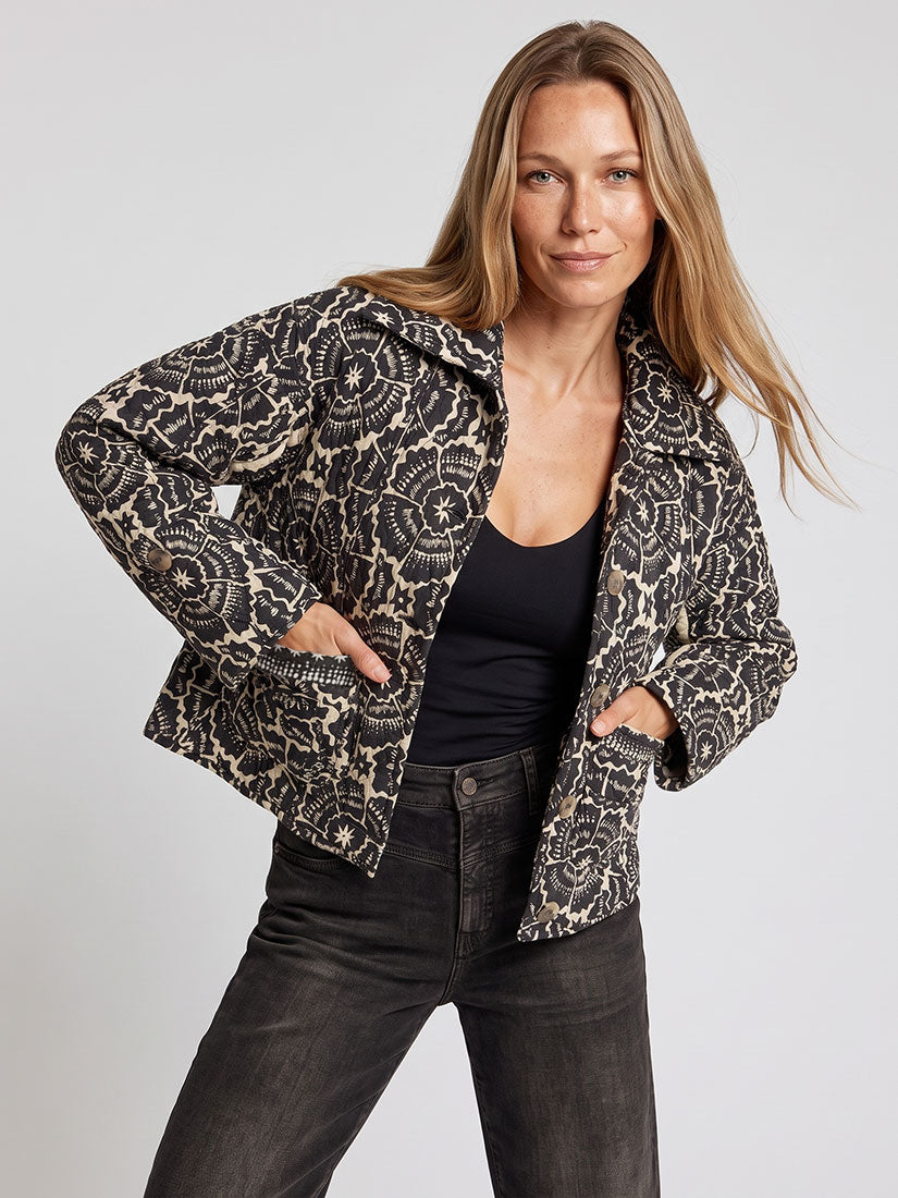A woman modeling a black and beige patterned jacket over a black top and dark jeans, standing against a light gray studio background.