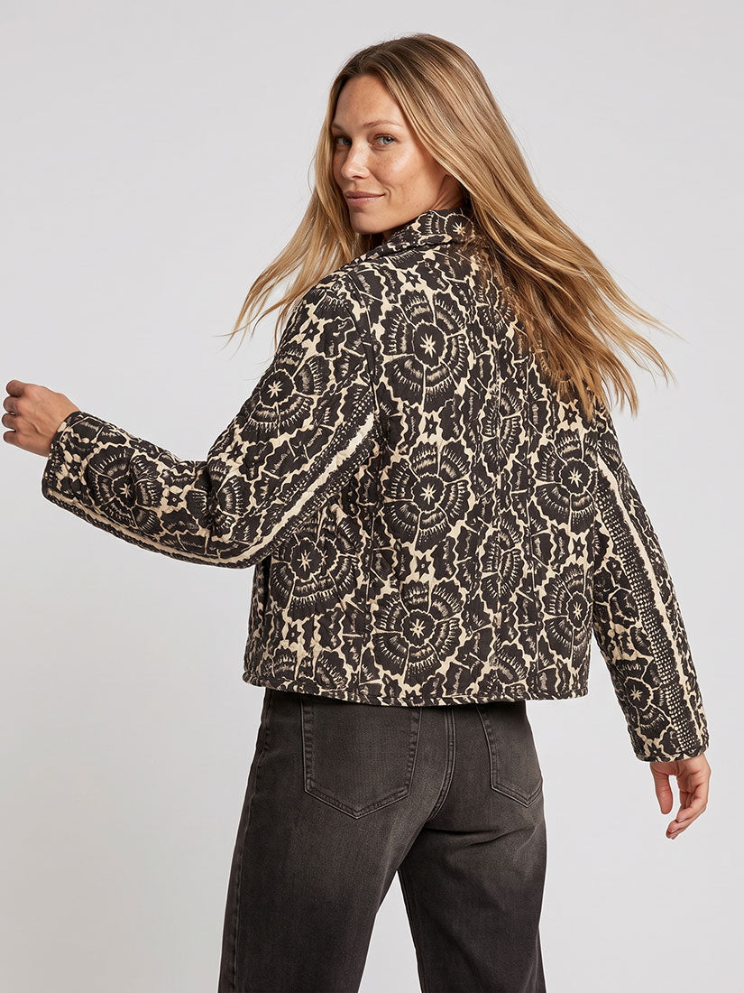 A woman wearing a cropped black and beige lace-patterned jacket, shown from a three-quarters back view against a light gray studio background.