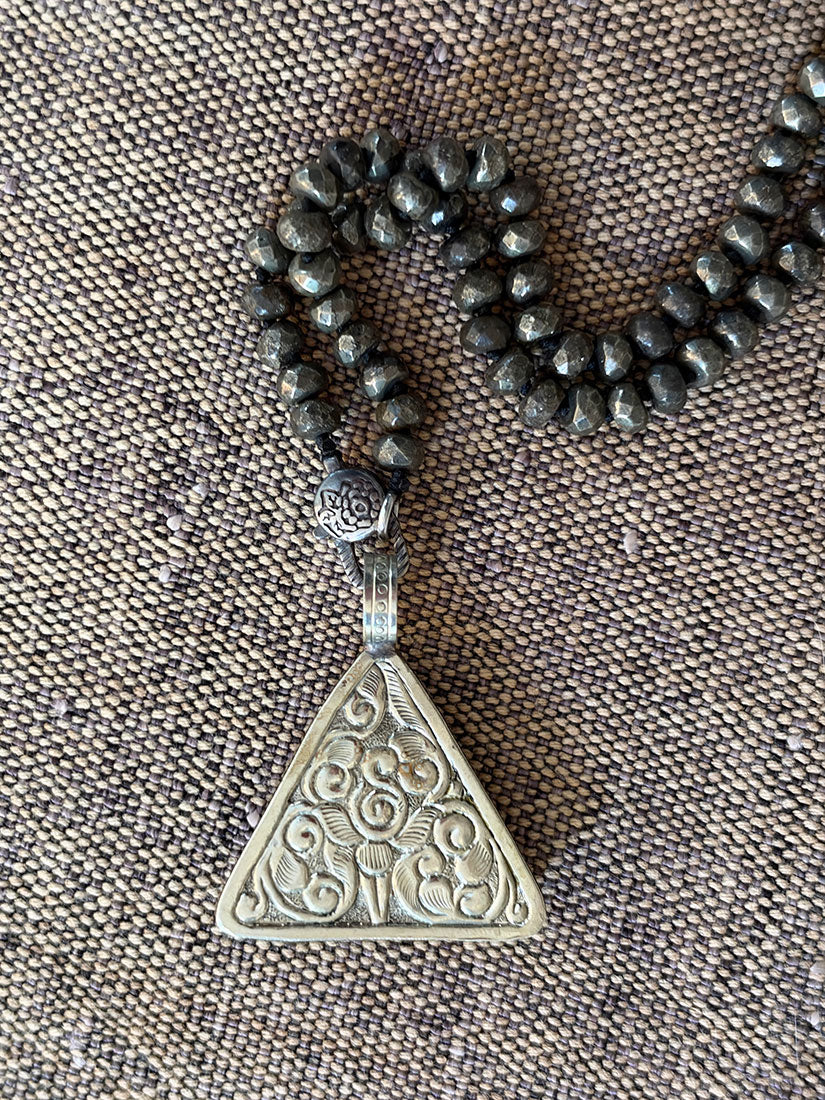 A silver-toned repousse triangular pendant necklace with intricate floral engravings, hanging from faceted pyrite beaded chain on a textured brown fabric background.